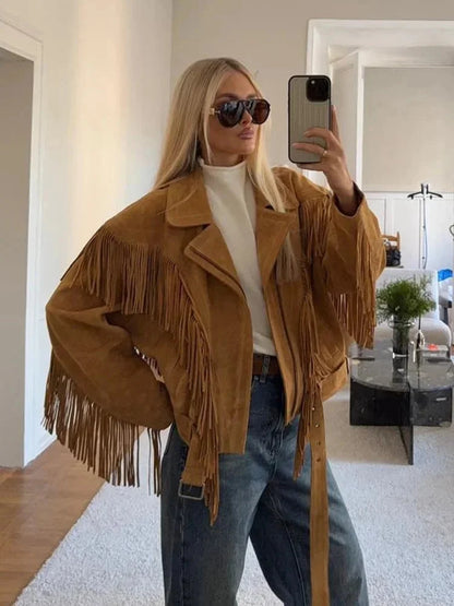 A woman wearing an oversized tan suede jacket featuring extensive fringe detailing along the chest and sleeves, styled with a white turtleneck and blue jeans.