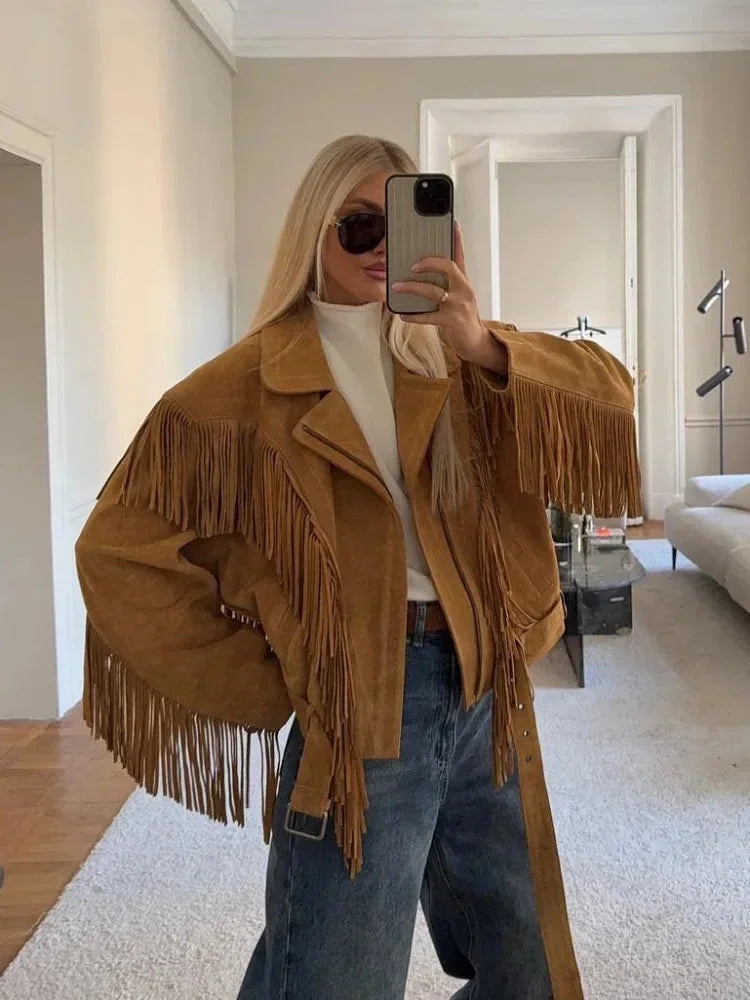 Brown - A woman wearing an oversized tan suede jacket featuring extensive fringe detailing along the chest and sleeves, styled with a white turtleneck and blue jeans.