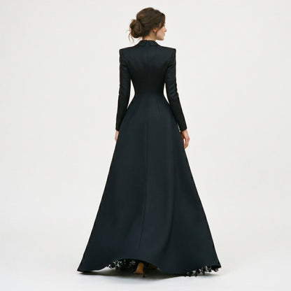 Black - A model poses in a structured black hybrid gown featuring a long-sleeve blazer-style bodice with peak lapels and a button-front closure, paired with a flowing floor-length skirt showcasing a sheer floral lace underlay and a high leg slit.