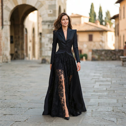 Black - A model poses in a structured black hybrid gown featuring a long-sleeve blazer-style bodice with peak lapels and a button-front closure, paired with a flowing floor-length skirt showcasing a sheer floral lace underlay and a high leg slit.