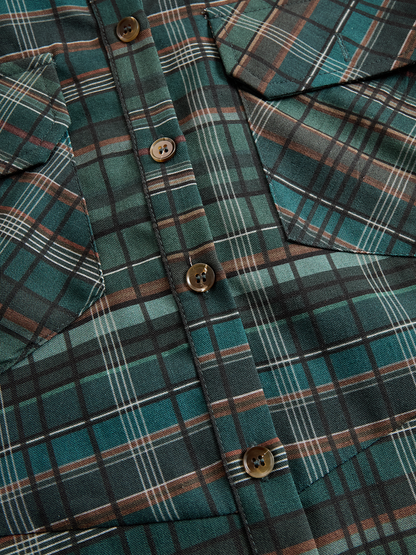 Black - A woman wearing a green and brown tartan plaid maxi shirt dress with a button-down front, long sleeves rolled up, and chest pockets, paired with brown leather cowboy boots against a textured grey wall.