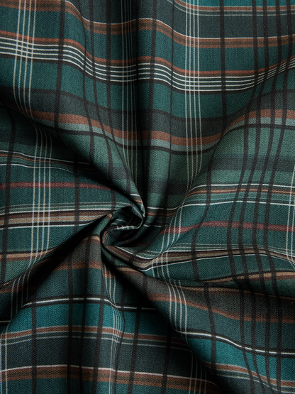 Black - A woman wearing a green and brown tartan plaid maxi shirt dress with a button-down front, long sleeves rolled up, and chest pockets, paired with brown leather cowboy boots against a textured grey wall.