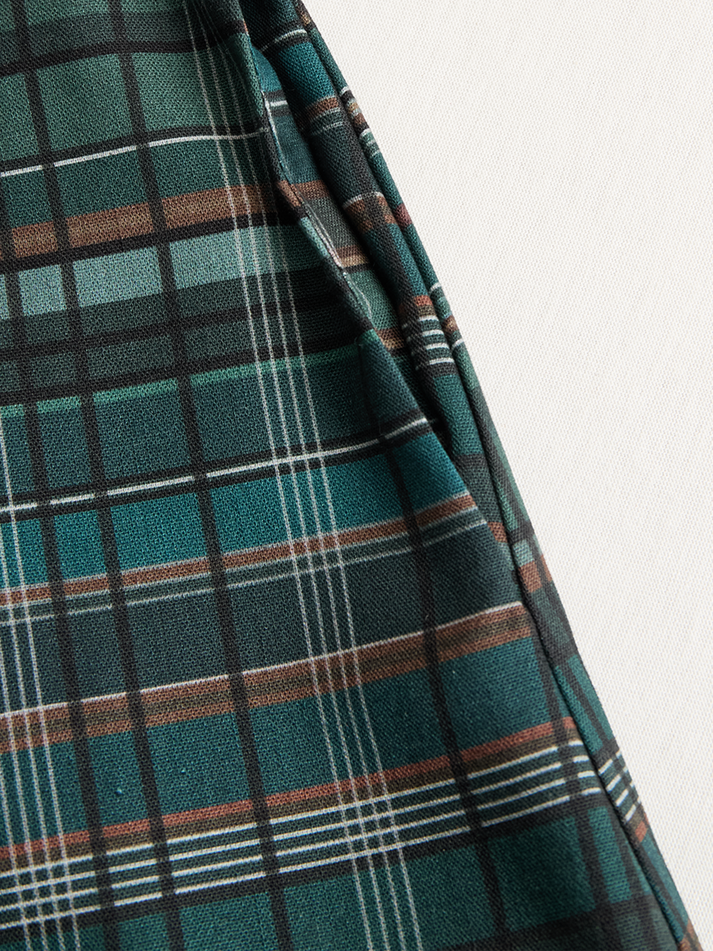 Black - A woman wearing a green and brown tartan plaid maxi shirt dress with a button-down front, long sleeves rolled up, and chest pockets, paired with brown leather cowboy boots against a textured grey wall.