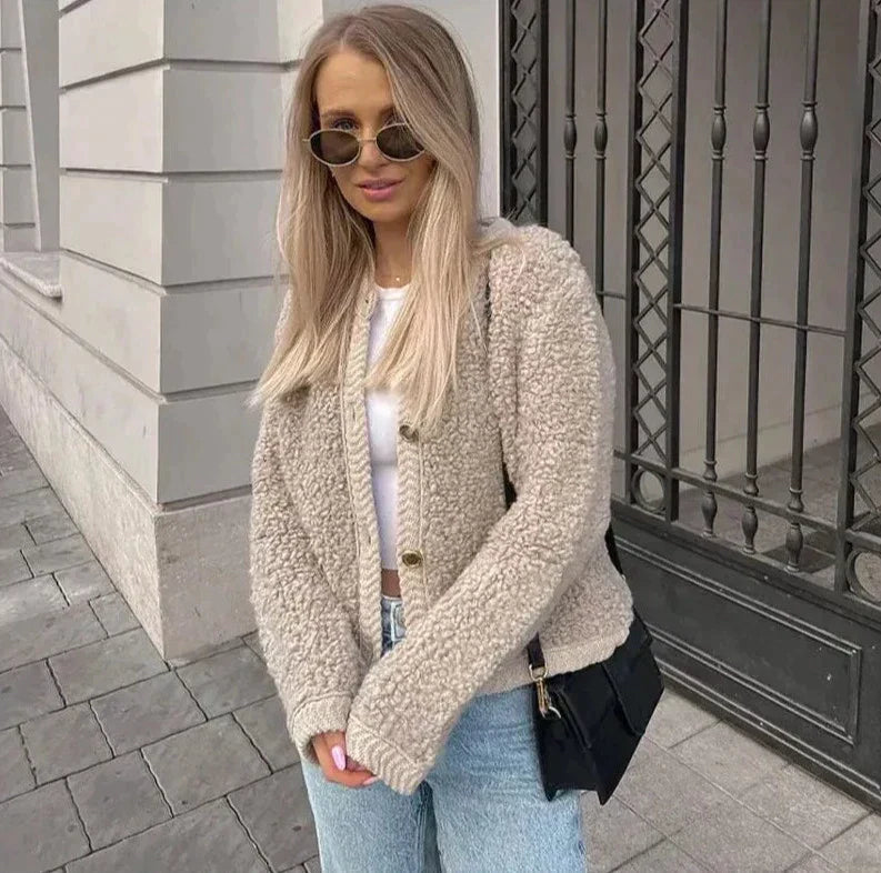 A woman wearing a beige, textured boucle knit cardigan with a button-front closure, ribbed trims, and gold-tone buttons, styled casually over a white top and light blue jeans.