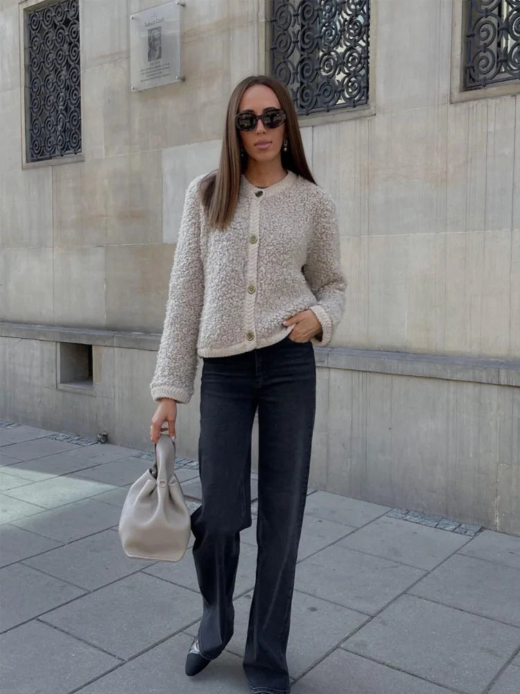 A woman wearing a beige, textured boucle knit cardigan with a button-front closure, ribbed trims, and gold-tone buttons, styled casually over a white top and light blue jeans.