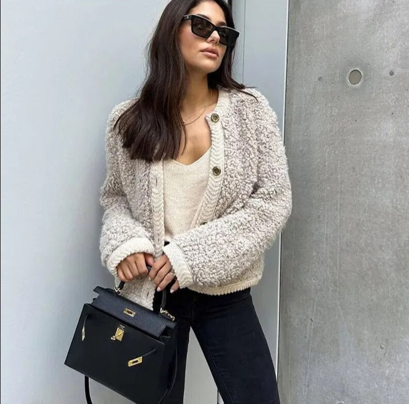 A woman wearing a beige, textured boucle knit cardigan with a button-front closure, ribbed trims, and gold-tone buttons, styled casually over a white top and light blue jeans.