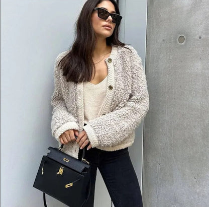 A woman wearing a beige, textured boucle knit cardigan with a button-front closure, ribbed trims, and gold-tone buttons, styled casually over a white top and light blue jeans.