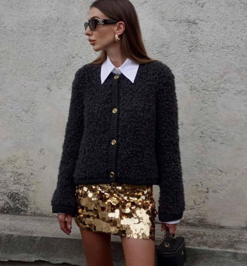 A woman wearing a beige, textured boucle knit cardigan with a button-front closure, ribbed trims, and gold-tone buttons, styled casually over a white top and light blue jeans.