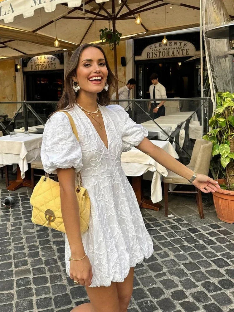 White - Women's white mini dress featuring a deep V-neckline, short puff sleeves, a structured bubble hem, and an all-over raised textured pattern, worn outdoors.