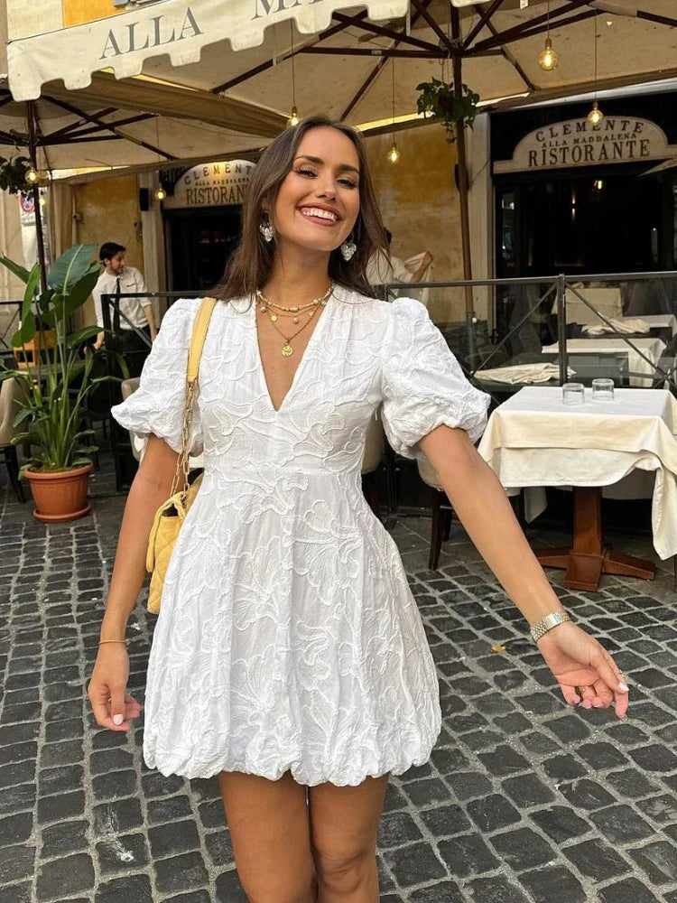 White - Women's white mini dress featuring a deep V-neckline, short puff sleeves, a structured bubble hem, and an all-over raised textured pattern, worn outdoors.