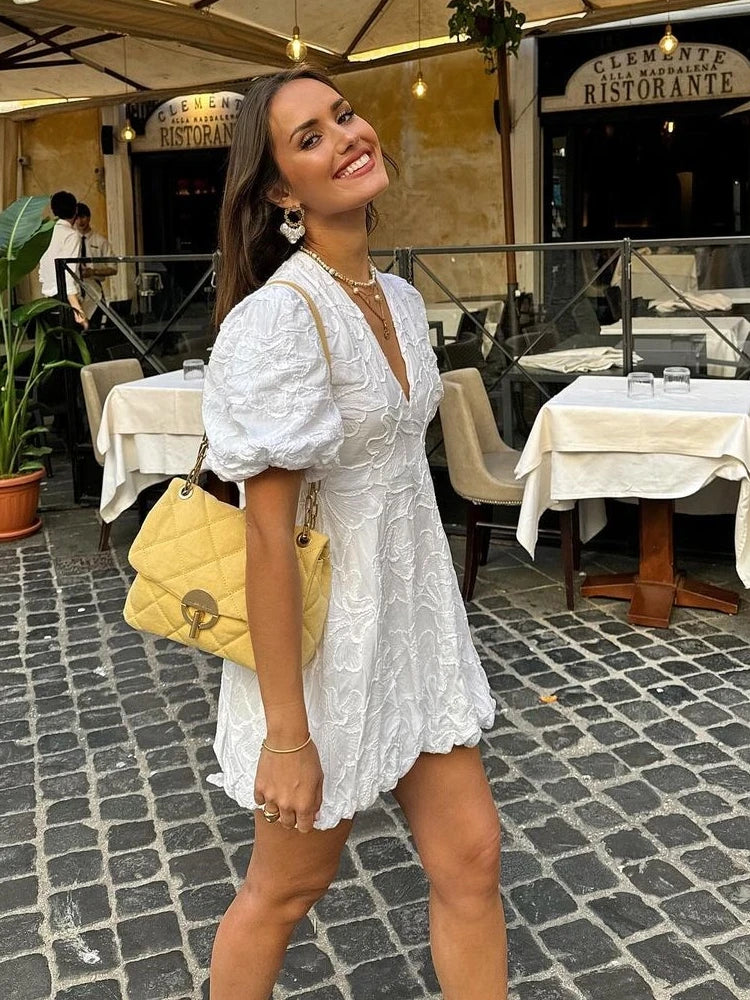 White - Women's white mini dress featuring a deep V-neckline, short puff sleeves, a structured bubble hem, and an all-over raised textured pattern, worn outdoors.