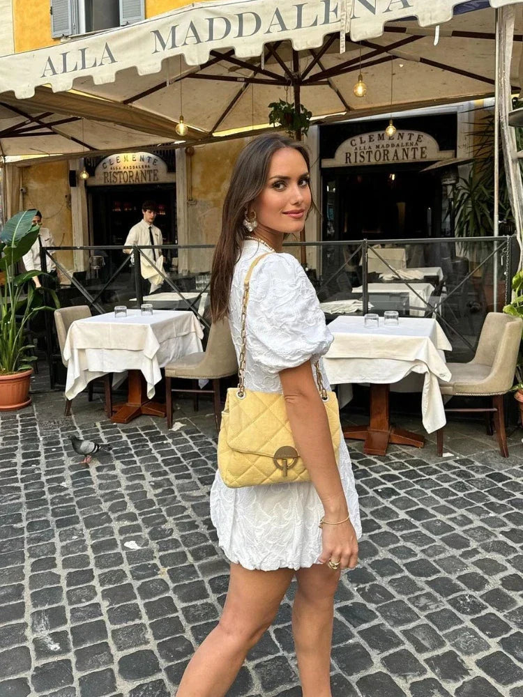 White - Women's white mini dress featuring a deep V-neckline, short puff sleeves, a structured bubble hem, and an all-over raised textured pattern, worn outdoors.