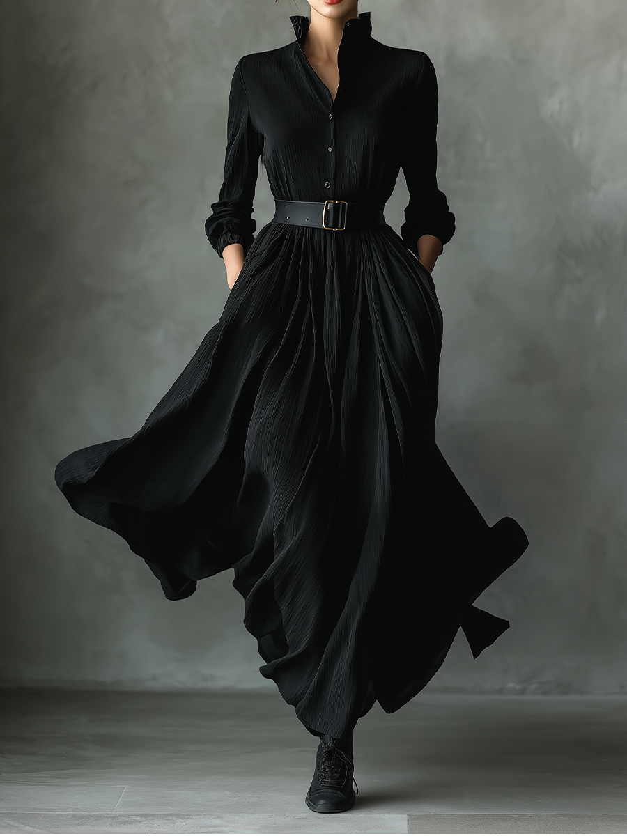 Black - A woman wearing a long-sleeved black textured crepe maxi dress with a high stand collar, button-up front, and a wide black belt with a gold buckle, showing a voluminous flowing skirt with hands in pockets.