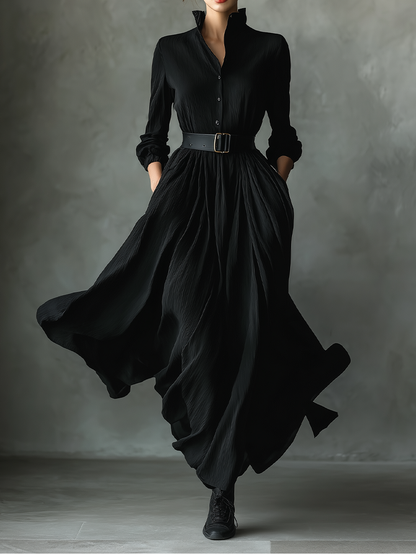 Black - A woman wearing a long-sleeved black textured crepe maxi dress with a high stand collar, button-up front, and a wide black belt with a gold buckle, showing a voluminous flowing skirt with hands in pockets.
