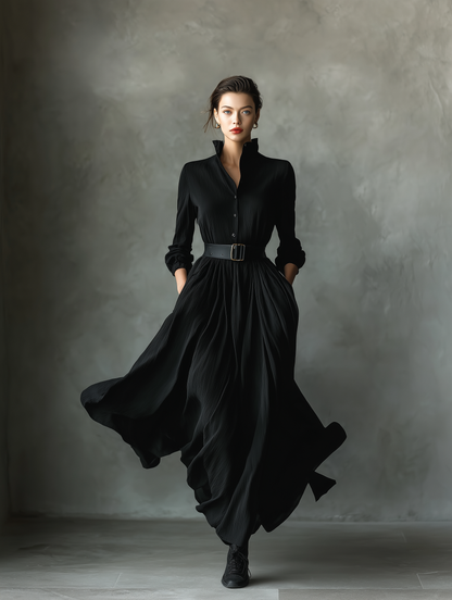 Black - A woman wearing a long-sleeved black textured crepe maxi dress with a high stand collar, button-up front, and a wide black belt with a gold buckle, showing a voluminous flowing skirt with hands in pockets.