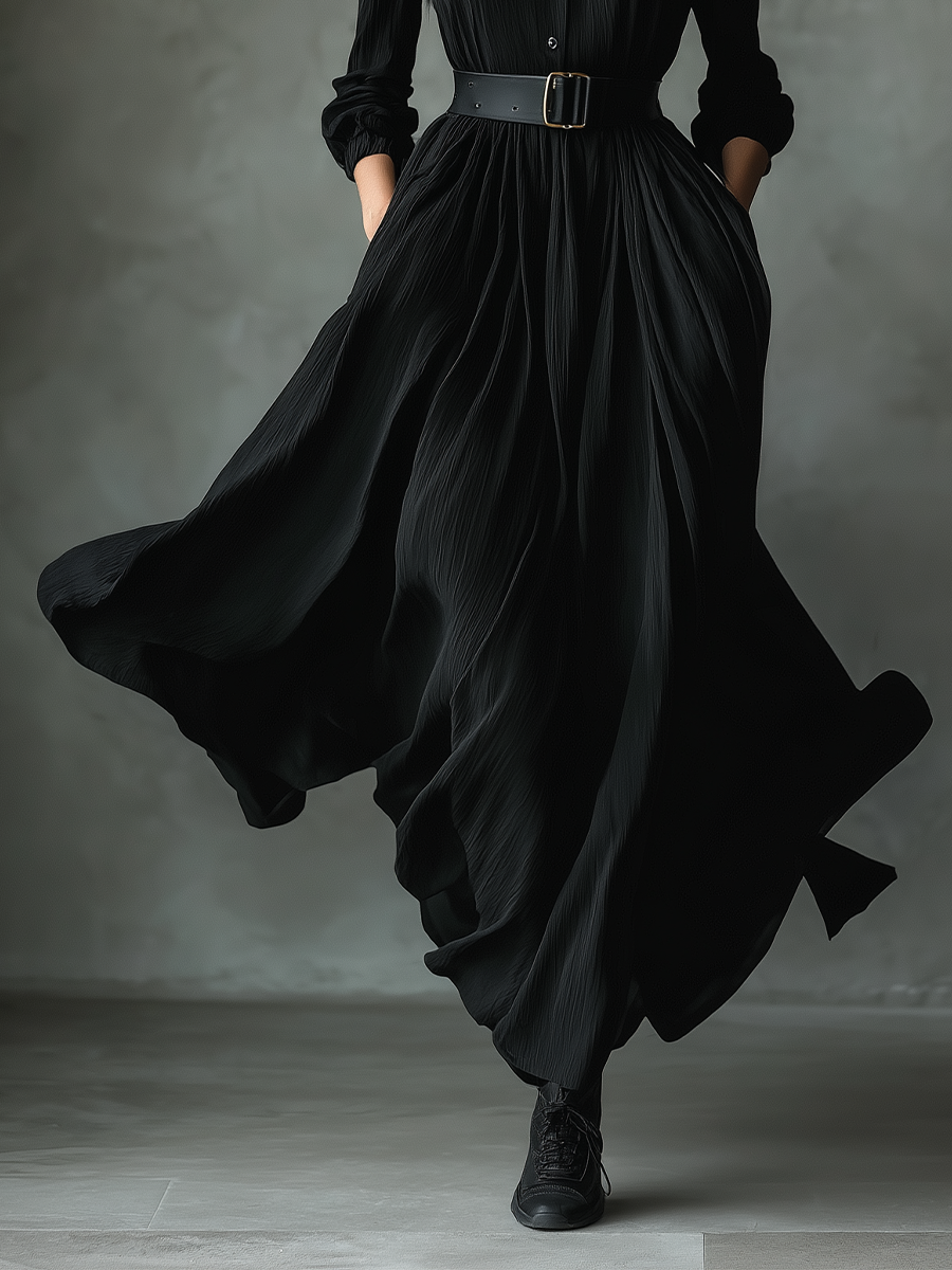 Black - A woman wearing a long-sleeved black textured crepe maxi dress with a high stand collar, button-up front, and a wide black belt with a gold buckle, showing a voluminous flowing skirt with hands in pockets.