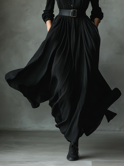 Black - A woman wearing a long-sleeved black textured crepe maxi dress with a high stand collar, button-up front, and a wide black belt with a gold buckle, showing a voluminous flowing skirt with hands in pockets.