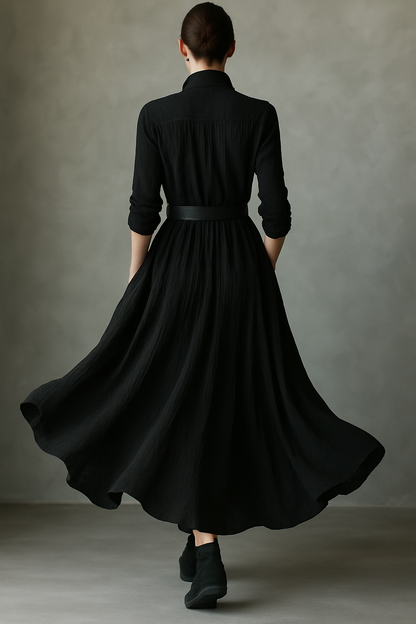 Black - A woman wearing a long-sleeved black textured crepe maxi dress with a high stand collar, button-up front, and a wide black belt with a gold buckle, showing a voluminous flowing skirt with hands in pockets.