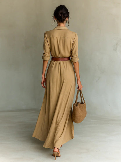 A woman wearing a sand-coloured, textured crepe maxi shirt dress with a plunging collared neckline, ruched waist detailing, and a draped wrap-effect skirt.