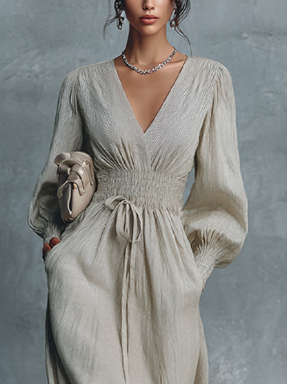Beige - A woman wearing a champagne-coloured, crinkle-textured crepe maxi dress featuring a plunging V-neckline, long balloon sleeves with shirred cuffs, a smocked waistband with a drawstring, and a flowing floor-length skirt.