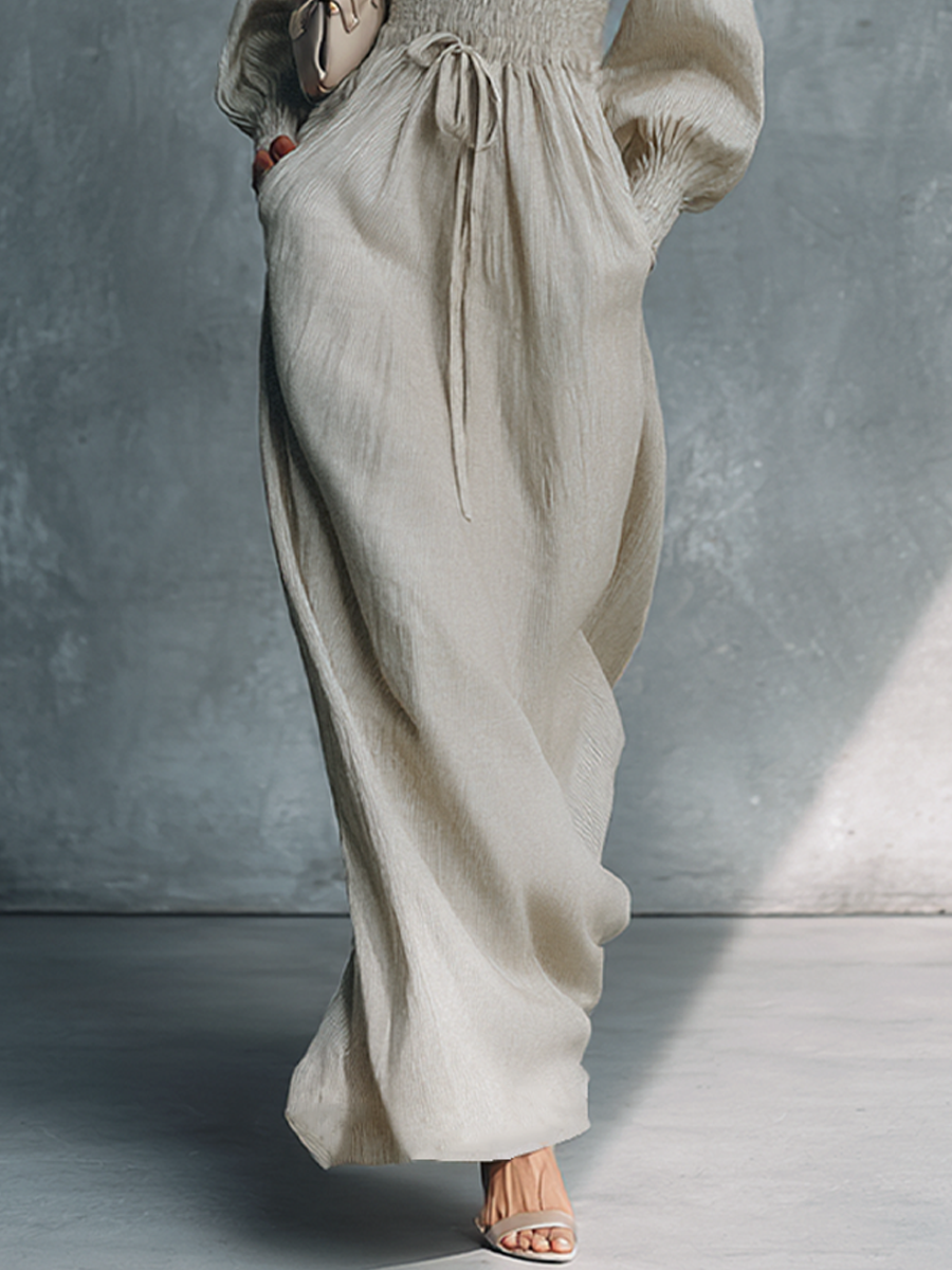 Beige - A woman wearing a champagne-coloured, crinkle-textured crepe maxi dress featuring a plunging V-neckline, long balloon sleeves with shirred cuffs, a smocked waistband with a drawstring, and a flowing floor-length skirt.