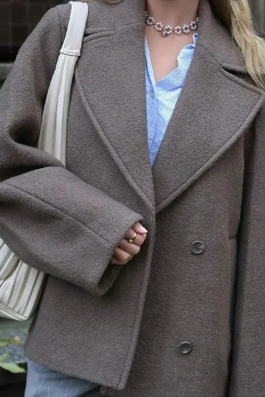Grey - A woman walking outdoors wearing a taupe-coloured, textured double-breasted short pea coat with wide notched lapels and an oversized fit, styled over a black top and high boots.
