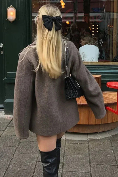Grey - A woman walking outdoors wearing a taupe-coloured, textured double-breasted short pea coat with wide notched lapels and an oversized fit, styled over a black top and high boots.