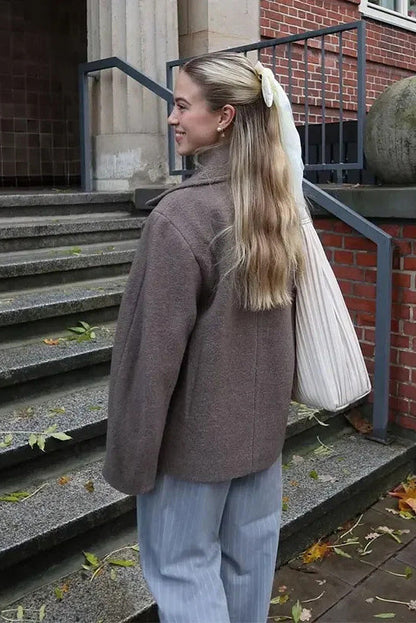 Grey - A woman walking outdoors wearing a taupe-coloured, textured double-breasted short pea coat with wide notched lapels and an oversized fit, styled over a black top and high boots.