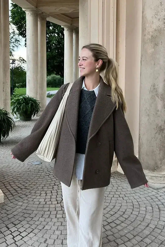 Grey - A woman walking outdoors wearing a taupe-coloured, textured double-breasted short pea coat with wide notched lapels and an oversized fit, styled over a black top and high boots.