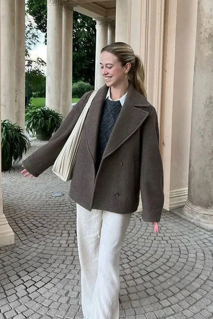 Grey - A woman walking outdoors wearing a taupe-coloured, textured double-breasted short pea coat with wide notched lapels and an oversized fit, styled over a black top and high boots.