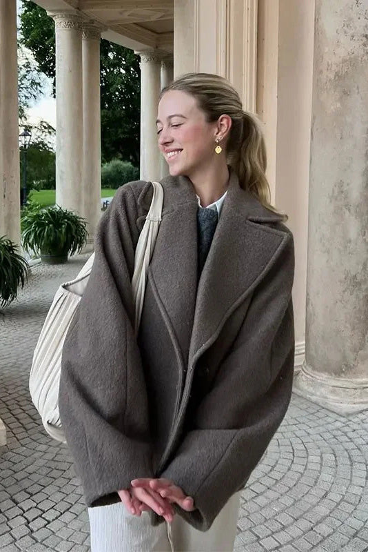 Grey - A woman walking outdoors wearing a taupe-coloured, textured double-breasted short pea coat with wide notched lapels and an oversized fit, styled over a black top and high boots.