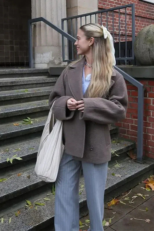 Grey - A woman walking outdoors wearing a taupe-coloured, textured double-breasted short pea coat with wide notched lapels and an oversized fit, styled over a black top and high boots.