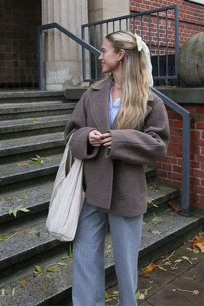 Grey - A woman walking outdoors wearing a taupe-coloured, textured double-breasted short pea coat with wide notched lapels and an oversized fit, styled over a black top and high boots.
