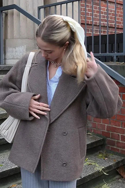 Grey - A woman walking outdoors wearing a taupe-coloured, textured double-breasted short pea coat with wide notched lapels and an oversized fit, styled over a black top and high boots.