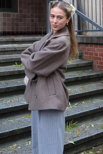 Grey - A woman walking outdoors wearing a taupe-coloured, textured double-breasted short pea coat with wide notched lapels and an oversized fit, styled over a black top and high boots.