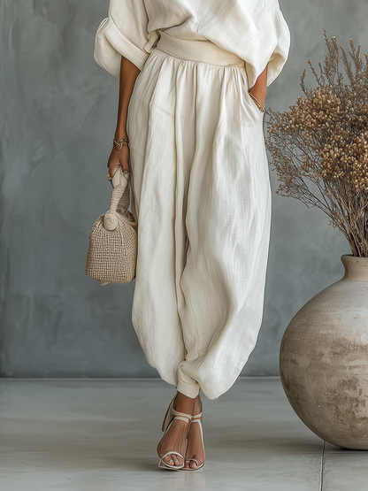 Off-white - A woman wearing an elegant cream-coloured, textured linen one-shoulder jumpsuit with a draped balloon sleeve, structured waistband, and voluminous tapered trousers.