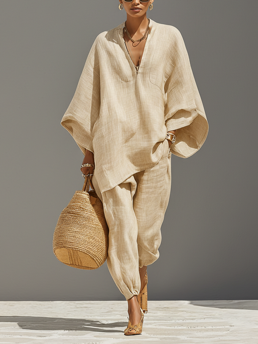 Neutral - A woman walking in a minimalist textured beige linen two-piece set featuring an oversized split-neck tunic with wide dolman sleeves and matching relaxed tapered trousers, accessorised with a woven straw tote bag.