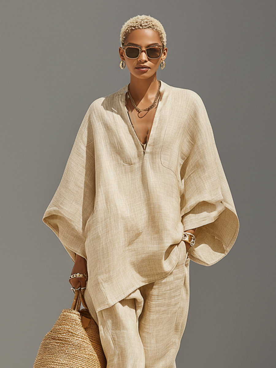 Neutral - A woman walking in a minimalist textured beige linen two-piece set featuring an oversized split-neck tunic with wide dolman sleeves and matching relaxed tapered trousers, accessorised with a woven straw tote bag.
