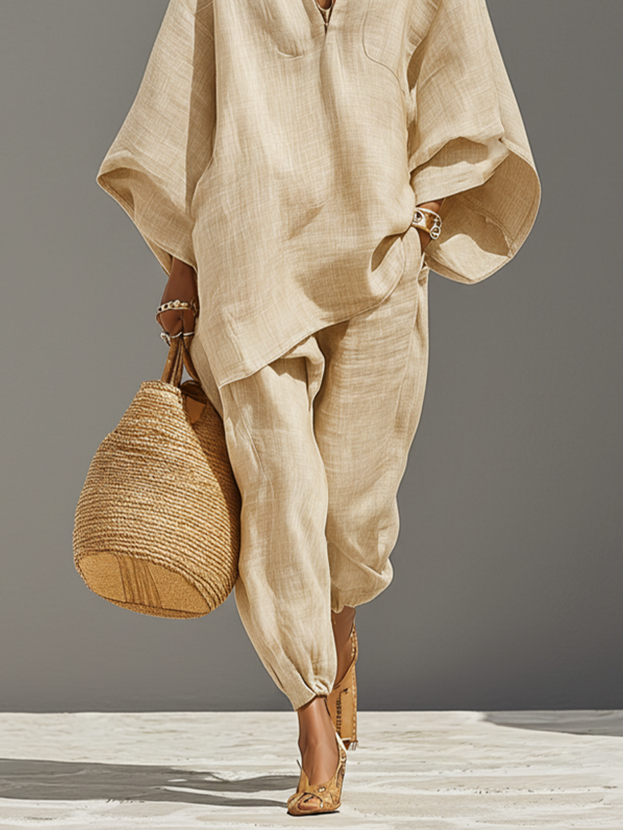 Neutral - A woman walking in a minimalist textured beige linen two-piece set featuring an oversized split-neck tunic with wide dolman sleeves and matching relaxed tapered trousers, accessorised with a woven straw tote bag.