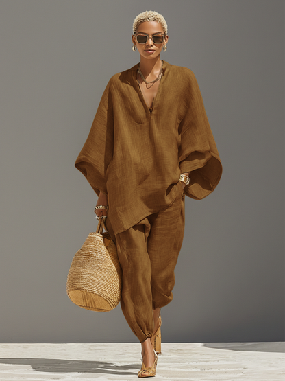 Tangerine - A woman walking in a minimalist textured beige linen two-piece set featuring an oversized split-neck tunic with wide dolman sleeves and matching relaxed tapered trousers, accessorised with a woven straw tote bag.