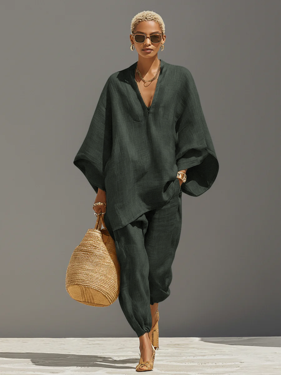 Grey - A woman walking in a minimalist textured beige linen two-piece set featuring an oversized split-neck tunic with wide dolman sleeves and matching relaxed tapered trousers, accessorised with a woven straw tote bag.