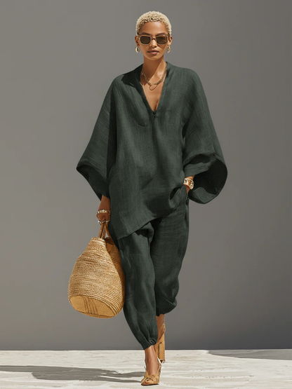 Grey - A woman walking in a minimalist textured beige linen two-piece set featuring an oversized split-neck tunic with wide dolman sleeves and matching relaxed tapered trousers, accessorised with a woven straw tote bag.