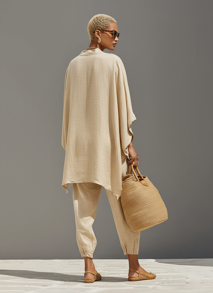 Grey - A woman walking in a minimalist textured beige linen two-piece set featuring an oversized split-neck tunic with wide dolman sleeves and matching relaxed tapered trousers, accessorised with a woven straw tote bag.