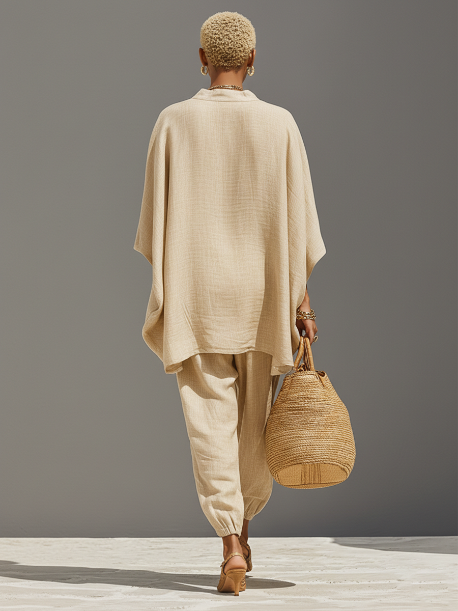 Grey - A woman walking in a minimalist textured beige linen two-piece set featuring an oversized split-neck tunic with wide dolman sleeves and matching relaxed tapered trousers, accessorised with a woven straw tote bag.