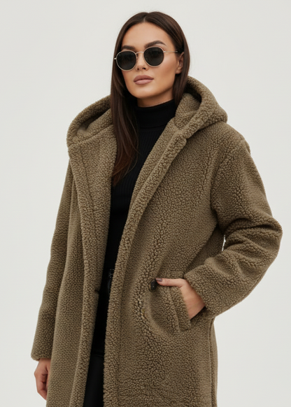 Khaki - A woman wearing a mid-length olive green textured teddy coat with a large hood, styled over a black turtleneck sweater and black sunglasses against a plain white background.