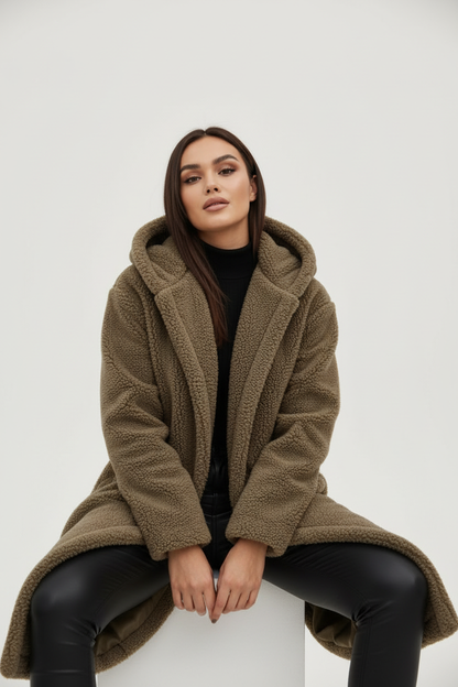 Khaki - A woman wearing a mid-length olive green textured teddy coat with a large hood, styled over a black turtleneck sweater and black sunglasses against a plain white background.