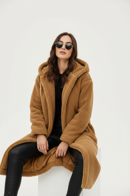 Camel - A woman wearing a mid-length olive green textured teddy coat with a large hood, styled over a black turtleneck sweater and black sunglasses against a plain white background.
