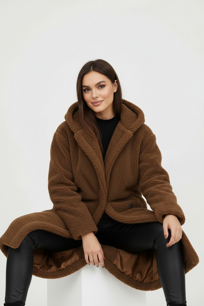 Brown - A woman wearing a mid-length olive green textured teddy coat with a large hood, styled over a black turtleneck sweater and black sunglasses against a plain white background.