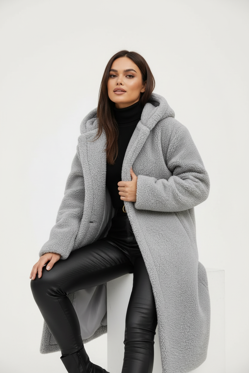 Grey - A woman wearing a mid-length olive green textured teddy coat with a large hood, styled over a black turtleneck sweater and black sunglasses against a plain white background.
