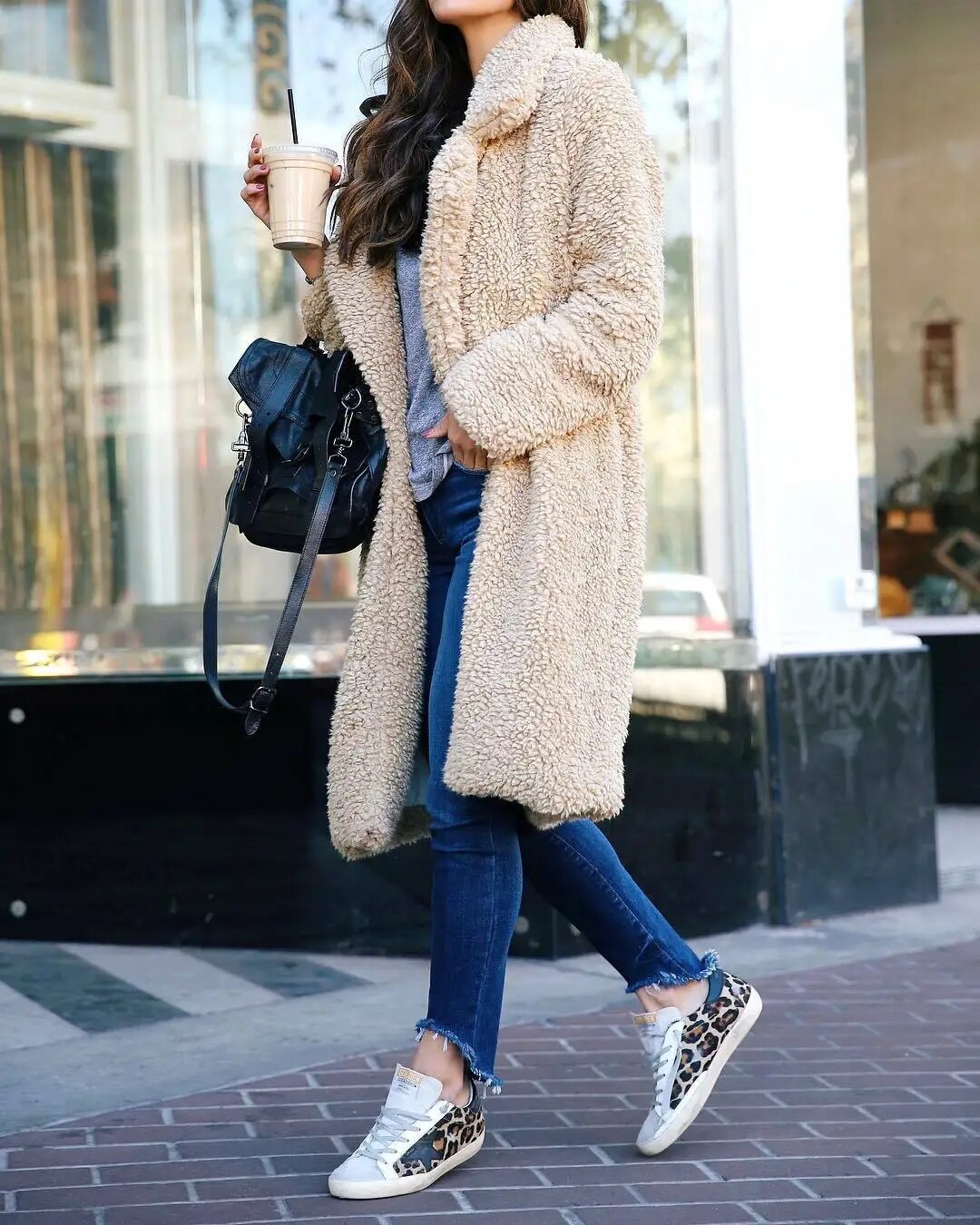 White - A woman walking on a city street wearing a knee-length, beige textured teddy coat with notch lapels, styled over a grey top and blue denim jeans.